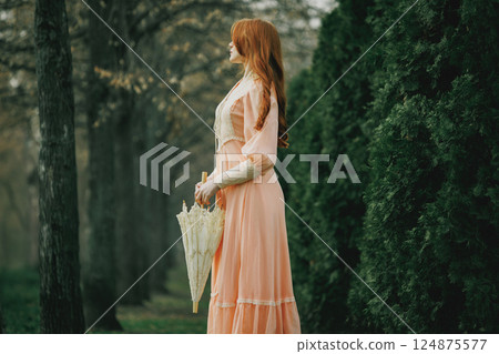 Young woman with auburn hair in delicate vintage dress stands still near tall firs, holding ivory umbrella, lost in silence. 124875577
