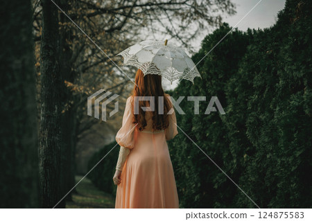 Red-haired woman in peach gown holds lace umbrella, moving between trees and shadows, evoking old-world charm and quiet longing. Red-haired woman in peach gown holds lace umbrella, moving between trees and shadows, evoking old-world charm and quiet longing. 124875583