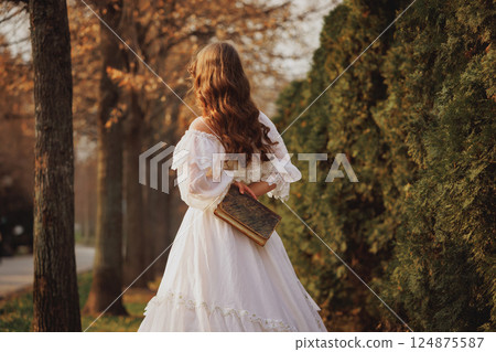 Girl holds vintage book behind her back while walking through autumn park in soft golden light, carrying echoes of past. Girl holds vintage book behind her back while walking through autumn park in soft golden light, carrying echoes of past. 124875587