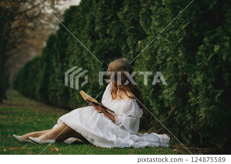 Woman in white dress sits on grass with antique book, facing away, surrounded by hedges and silence. Suitable for book or film cover. 124875589
