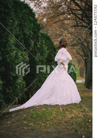 Girl in historical white dress stands still beside green cypress hedge, surrounded by bare trees, lost in thoughts. Suitable for book or film cover Girl in historical white dress stands still beside green cypress hedge, surrounded by bare trees, lost in thoughts. Suitable for book or film cover 124875592