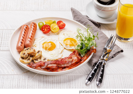 A classic breakfast plate featuring sunny-side-up eggs, crispy bacon, sausages, and fresh tomatoes 124877022