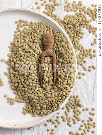 Plate full of dried raw green lentils with a wooden scoop on white table top view. Vegetarian food Plate full of dried raw green lentils with a wooden scoop on white table top view. Vegetarian food 124877986