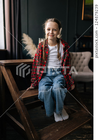 Full length vertical portrait of smiling preteen with braided hair wearing plaid shirt, sitting on ladder against dark wall. Cheerful child girl posting in casual clothes smiling looking at camera. 124879239