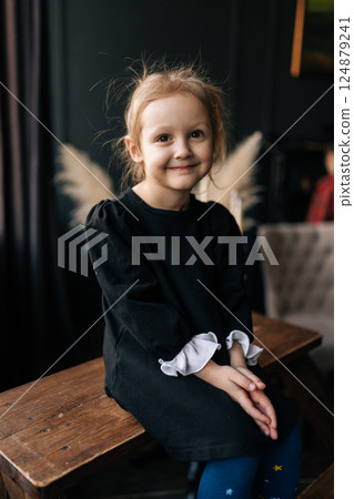 Vertical portrait of pretty young girl wearing ruffled top and blue tights sitting on wooden bench, hands gently clasped, radiating pure childhood innocence with soft smile, looking at camera. 124879241