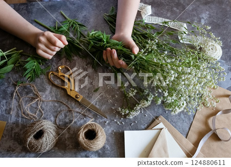 Florist at work: a pretty woman prepares a summer bouquet of white gypsophila on a gray desk. Kraft paper, scissors, greeting envelope on the table. View from above. The composition is flat. Florist at work: a pretty woman prepares a summer bouquet of white gypsophila on a gray desk. Kraft paper, scissors, greeting envelope on the table. View from above. The composition is flat. 124880611