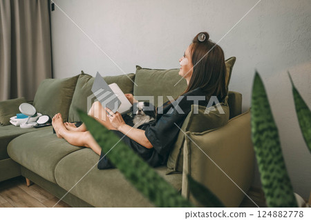 woman with eye patches relaxing on couch, reading a book, and petting dog in a cozy living room, concept of self-care and pet care 124882778