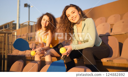Portrait of young women adults posing with pickleball paddle and ball on stadium chairs 124882892