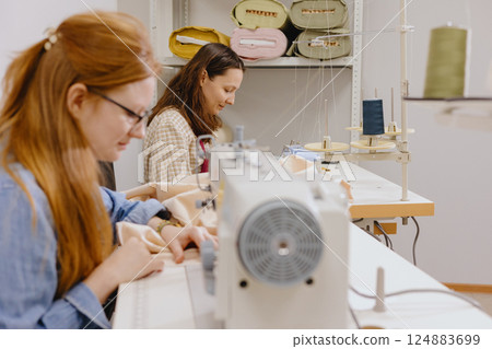 Two female seamstresses in the workshop at the factory are working at tables on sewing machines. Two female seamstresses in the workshop at the factory are working at tables on sewing machines. 124883699