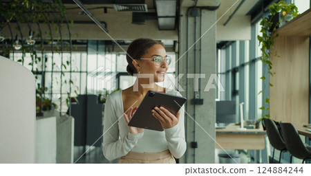 Happy young woman in stylish outfit using a digital tablet in a modern office with plants and natural light through large glass windows 124884244