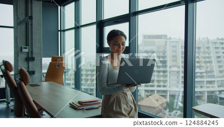 A young woman holding a laptop and working in a modern office with large windows and a panoramic city view, symbolizing professionalism and focus A young woman holding a laptop and working in a modern office with large windows and a panoramic city view, symbolizing professionalism and focus 124884245