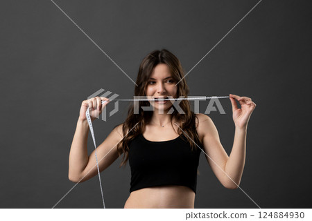 Angry brunette woman in black t-shirt holding a centimeter tape in a mouth with a grey background. Sport, fitness and diet. 124884930