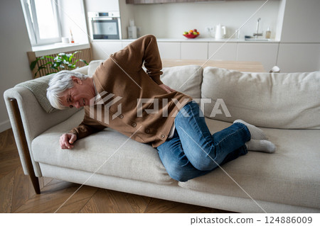 Elderly man struggles to rise from couch, hunched with back pain, spinal osteochondrosis, cystitis 124886009