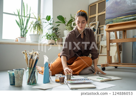 Serious thoughtful female artist paint watercolor in sketchbook on floor in workshop, artwork studio 124886053
