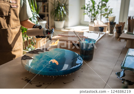 Woman artist hands carefully removes bubbles using gas torch on round epoxy resin piece in studio 124886219
