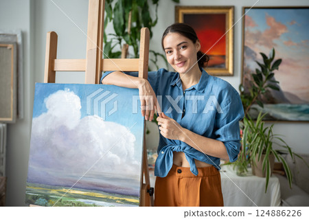 Portrait proud happy woman artist with new painting of sky and nature in workshop, art studio Portrait proud happy woman artist with new painting of sky and nature in workshop, art studio 124886226