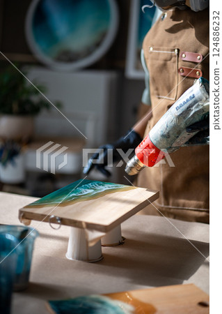 Artist hands use hot air gun to clear excess epoxy from wave painting on board. Handmade painting 124886232