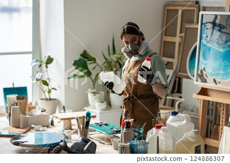 Focused craftswoman in respirator handles reagents preparing epoxy solution for artwork in studio Focused craftswoman in respirator handles reagents preparing epoxy solution for artwork in studio 124886307