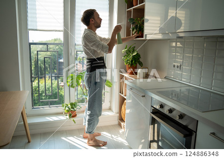 Man wearing spine corset for fixing lower back spraying plants on kitchen at home in sick leave. Man wearing spine corset for fixing lower back spraying plants on kitchen at home in sick leave. 124886348