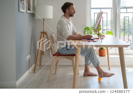 Man freelancer working on computer at home doing recovery foot exercise using massage silicone ball. 124886377