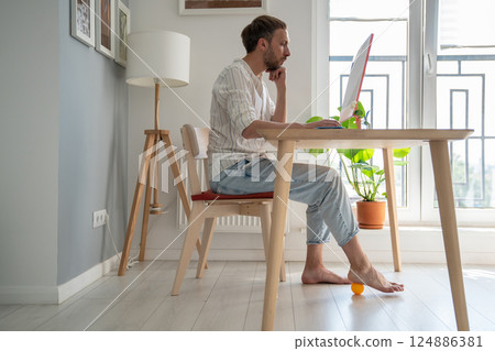 Man freelancer working on computer at home doing recovery foot exercise using massage silicone ball. 124886381