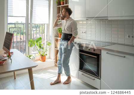 Man in back support corset on lower back to treatment of hernia on kitchen drinking water. Man in back support corset on lower back to treatment of hernia on kitchen drinking water. 124886445