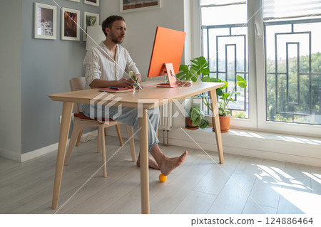 Man IT freelancer working on computer at home doing foot exercise using massage ball. Man IT freelancer working on computer at home doing foot exercise using massage ball. 124886464