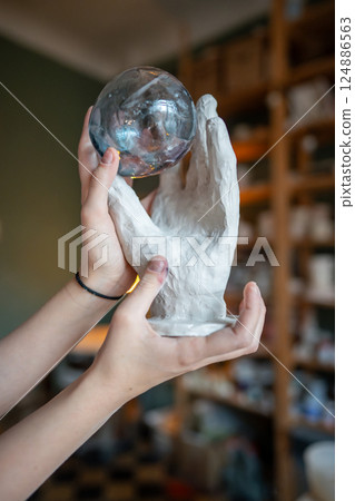 Glass ball rests in handcrafted clay hand decor in ceramist hands. Glassmaking and pottery as art Glass ball rests in handcrafted clay hand decor in ceramist hands. Glassmaking and pottery as art 124886563