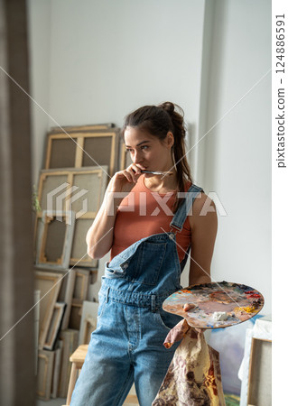 Inspired woman artist holds palette, paintbrush pensive looks to canvas, think of idea in art studio 124886591