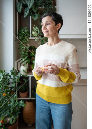 Relaxed cheerful woman peel mandarin tangerine, looking out kitchen window. Slow life tranquility 124886601
