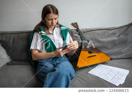 Woman musician searches for notes and surfs internet in smartphone while notes, balalaika on couch 124886615
