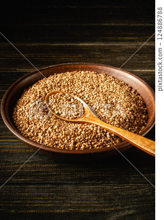 A wooden bowl filled with small brown buckwheat seeds sits on a dark wooden surface. Inside lies a wooden spoon, indicating preparation for cooking or seasoning A wooden bowl filled with small brown buckwheat seeds sits on a dark wooden surface. Inside lies a wooden spoon, indicating preparation for cooking or seasoning 124886788