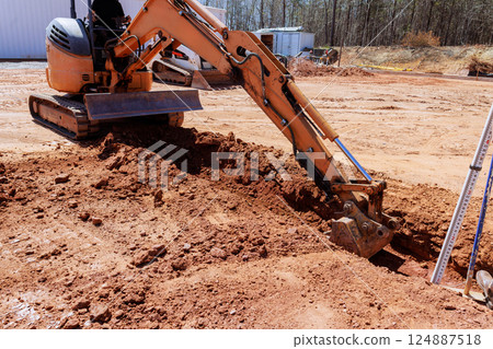Construction worker uses an excavator to dig trench in dirt field during works construction 124887518