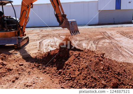 Excavator digs into ground, moving soil during construction activity at site under works construction 124887521