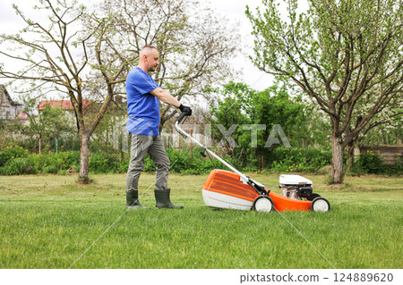 Man in protective gloves, gardener is mowing bright lush green grass lawn using modern gasoline cordless lawnmower at own backyard. Seasonal landscaping design work. Gardening work tools. Man in protective gloves, gardener is mowing bright lush green grass lawn using modern gasoline cordless lawnmower at own backyard. Seasonal landscaping design work. Gardening work tools. 124889620