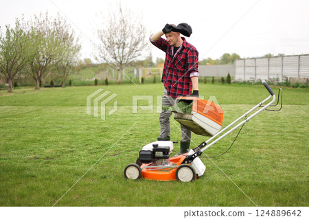 Tired man in casual clothes holds bag with cut grass of lawn mower after mowing lawn with at own backyard. Husband takes care of garden. Modern gasoline garden equipment. Landscaping work at home 124889642