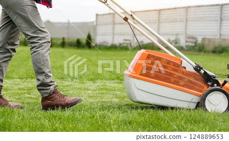 Unrecognizable professional gardener in protective apparel is mowing green grass lawn using modern gasoline cordless lawnmower at backyard. Seasonal landscaping design work. Gardening work tools. Unrecognizable professional gardener in protective apparel is mowing green grass lawn using modern gasoline cordless lawnmower at backyard. Seasonal landscaping design work. Gardening work tools. 124889653