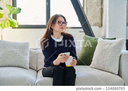 Relaxed mature woman with smartphone cup of drink sitting on sofa at home Relaxed mature woman with smartphone cup of drink sitting on sofa at home 124889703