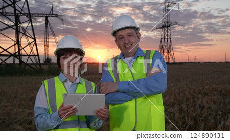 Man and woman engineers with tablet smile to camera against electric power generation plant in sunset field. Engineers work together at power distribution substation at twilight time closeup 124890431