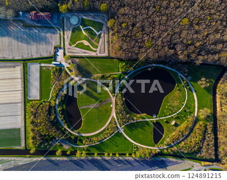 Aerial view of a uniquely designed park with circular paths and ponds, surrounded by colorful agricultural fields and bordered by a dense forest. A striking mix of nature and geometry. 124891215