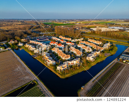 Aerial view of a peaceful suburban neighborhood surrounded by fields and canals. Neat rows of houses with red roofs reflect in the water, set against a backdrop of farmland and open skies. 124891224
