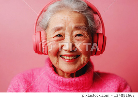 An Asian senior woman, smiling joyfully with headphones, enjoying the pleasure of music or studying, set against a pink-colored background, embodying the vibrant enjoyment of life in her later years 124891662