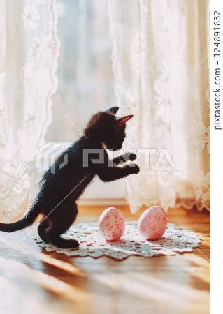 Small black kitten playing with Easter eggs on wooden table near window. Soft natural light, shadows 124891832