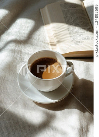 Warm coffee cup beside an open book on a sunlit table with leafy shadows 124893699