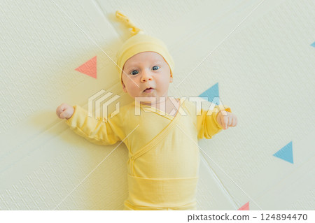 Adorable baby in a yellow hat lying on a play mat. Cozy newborn moment with soft textures and bright colors. Infant development, sensory play, and early childhood concept Adorable baby in a yellow hat lying on a play mat. Cozy newborn moment with soft textures and bright colors. Infant development, sensory play, and early childhood concept 124894470