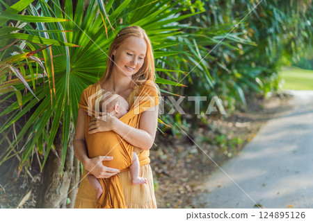 Mother holding her little baby in a yellow sling in the park. Warm and loving family moment. Babywearing, parenting, and mother-child bonding concept 124895126