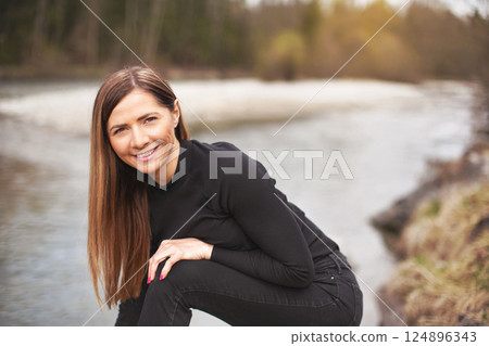 Portrait of young woman wearing black jeans and top, leaning, her long hair falling down, smiling. Blurred river in background Portrait of young woman wearing black jeans and top, leaning, her long hair falling down, smiling. Blurred river in background 124896343
