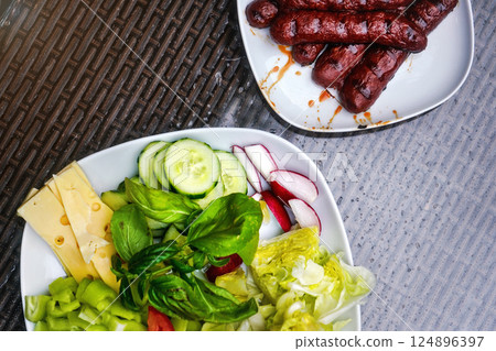 Two white plates, one with grilled bratwursts / frankfurters, other with salad - cucumbers, radish, basil, pepper and cheese. View from above 124896397