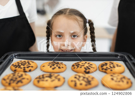 Caucasian young girl with braided hair, wearing apron, smiling, displaying baked chocolate chip cookies, surrounded by family members engaged in baking activity. 124902806