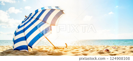 Striped blue and white umbrella casting shadow on sandy beach near seashells with sunlit ocean and clear sky in background, conveying a tranquil summer day at the coast 124908108